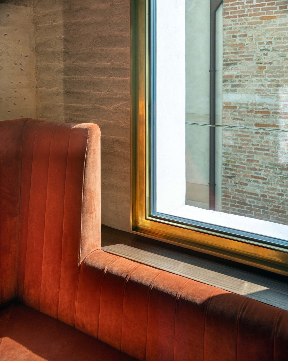 Detail of a burnished bronze window frame against exposed brick, with a velvet banquette below