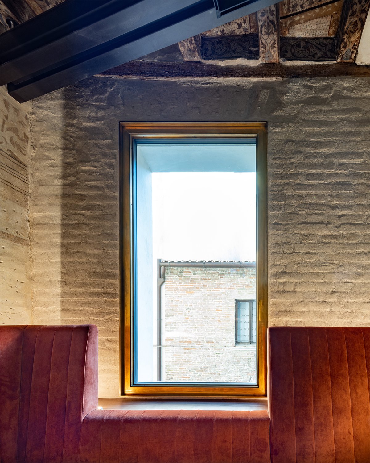 A tall bronze-framed window set into a rough plaster wall beneath a beamed ceiling